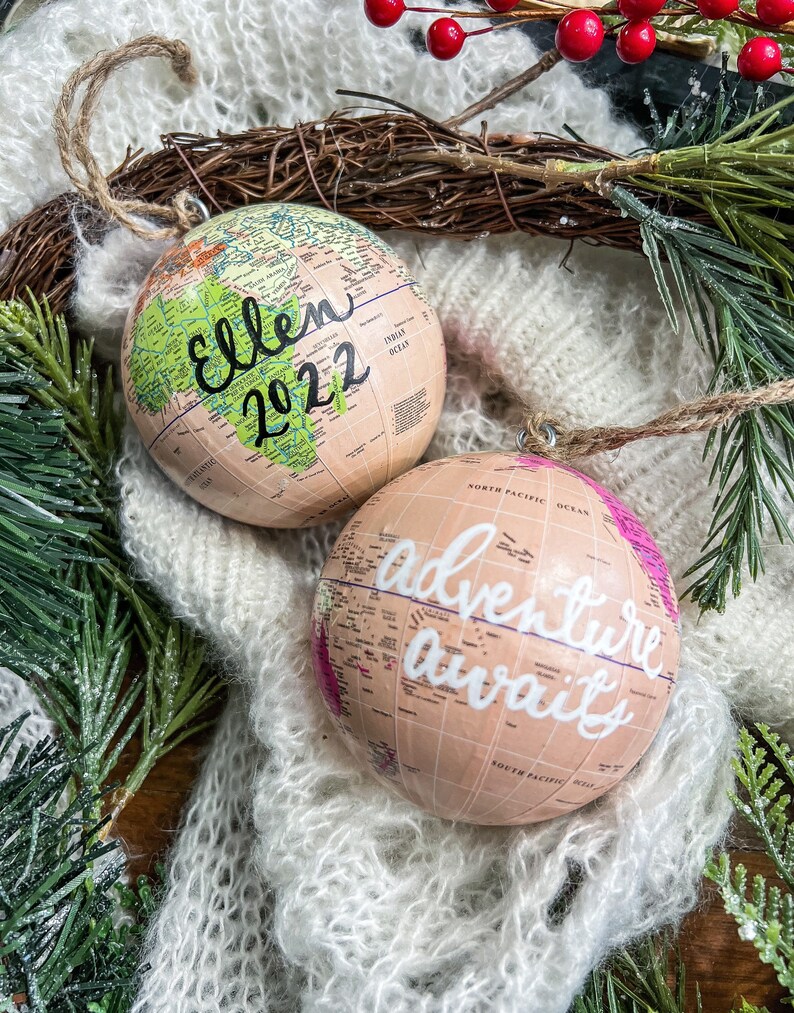 May include: Two globe-shaped Christmas ornaments. One is green and beige with the text "Ellen 2022" written on it. The other is pink and beige with the text "Adventure awaits" written on it.