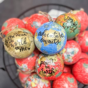 May include: A collection of globe ornaments with handwritten messages. The ornaments are in various colors, including red, gold, blue, and green. The messages include "You mean the world to me", "Adventure awaits", "Joy to the world", and "Peace on earth".