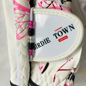 May include: White golf glove with a pink and black floral pattern. The glove has a white patch with the text "Birdie Town Jupiter" on it. A beaded bracelet is attached to the glove.