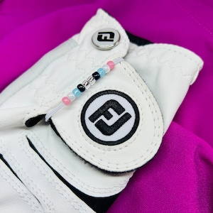 May include: White golf glove with a black and white logo on the back of the hand. A white elastic bracelet with pink, blue, and silver beads is attached to the glove.