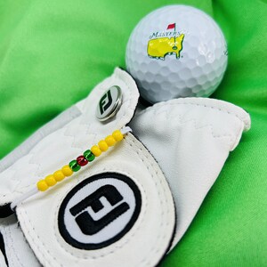 May include: White golf glove with a black "F" logo and a yellow, red, and green beaded strap. A white golf ball with the Masters Tournament logo is in the background.