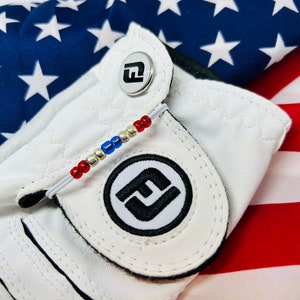 May include: White golf glove with a red, white, and blue beaded bracelet and a black and white logo on the back of the hand. The glove is on top of an American flag.