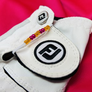 May include: White golf glove with a black and white logo and a pink and gold beaded strap. The glove has a snap closure.