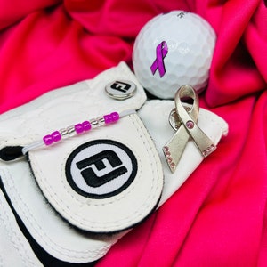 Puede incluir: Guante de golf blanco con una pulsera de cuentas rosa y blanca, una bola de golf blanca con una cinta rosa y un broche de cinta plateado con diamantes de imitación rosas.
