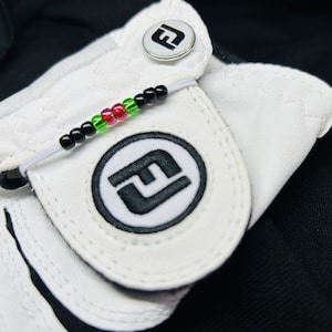 May include: White golf glove with a black and white logo and a black, green, and red beaded bracelet attached to the glove.