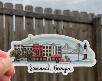 May include: A sticker featuring a colorful illustration of Savannah, Georgia's skyline. The design includes buildings, a bridge, a lighthouse, and a riverboat. The text "Savannah, Georgia" is written in a cursive font.