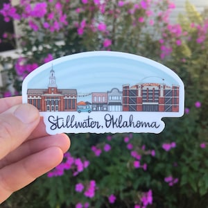 May include: A sticker featuring a colorful illustration of the Stillwater, Oklahoma skyline. The design includes buildings, a blue sky, and the text "Stillwater, Oklahoma" in a handwritten font. The sticker is held against a backdrop of green foliage and pink flowers.