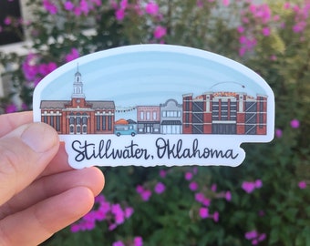 May include: A sticker featuring a colorful illustration of the Stillwater, Oklahoma skyline. The design includes buildings, a blue sky, and the text "Stillwater, Oklahoma" in a handwritten font. The sticker is held against a backdrop of green foliage and pink flowers.