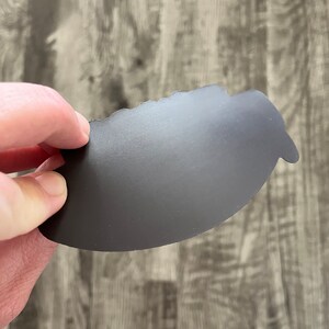 May include: A dark gray, irregularly shaped magnet is held against a wooden surface. The magnet has a matte finish and a slightly textured surface. The edges are uneven, giving it a unique, handcrafted appearance. The background is a light gray wood grain.