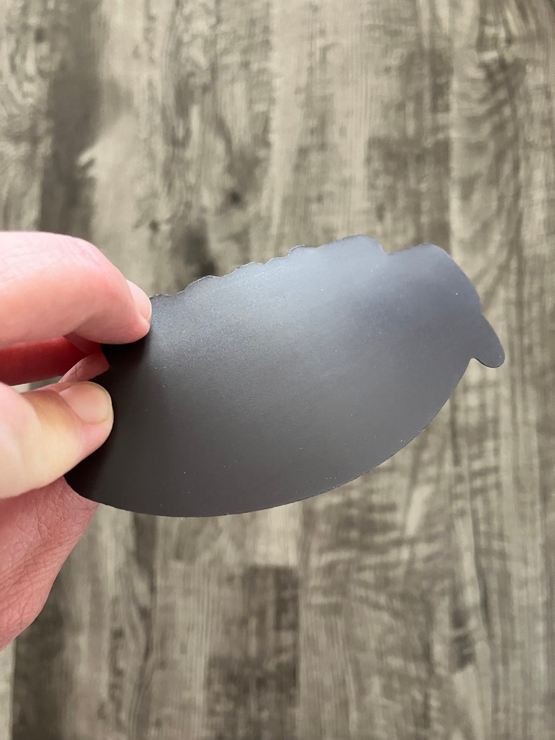 May include: A dark gray, irregularly shaped magnet is held against a wooden surface. The magnet has a matte finish and a slightly textured surface. The edges are uneven, giving it a unique, handcrafted appearance. The background is a light gray wood grain.