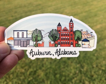 May include: A sticker featuring an illustrated skyline of Auburn, Alabama, with buildings, trees, and a stadium. The sticker has a white border and the text "Auburn, Alabama" in a handwritten font.
