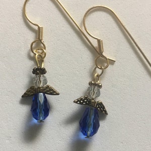 Op de afbeelding: Een paar gouden oorhangers met een blauwe kristallen traan en een klein gouden engelbedeltje met heldere kristallen accenten.