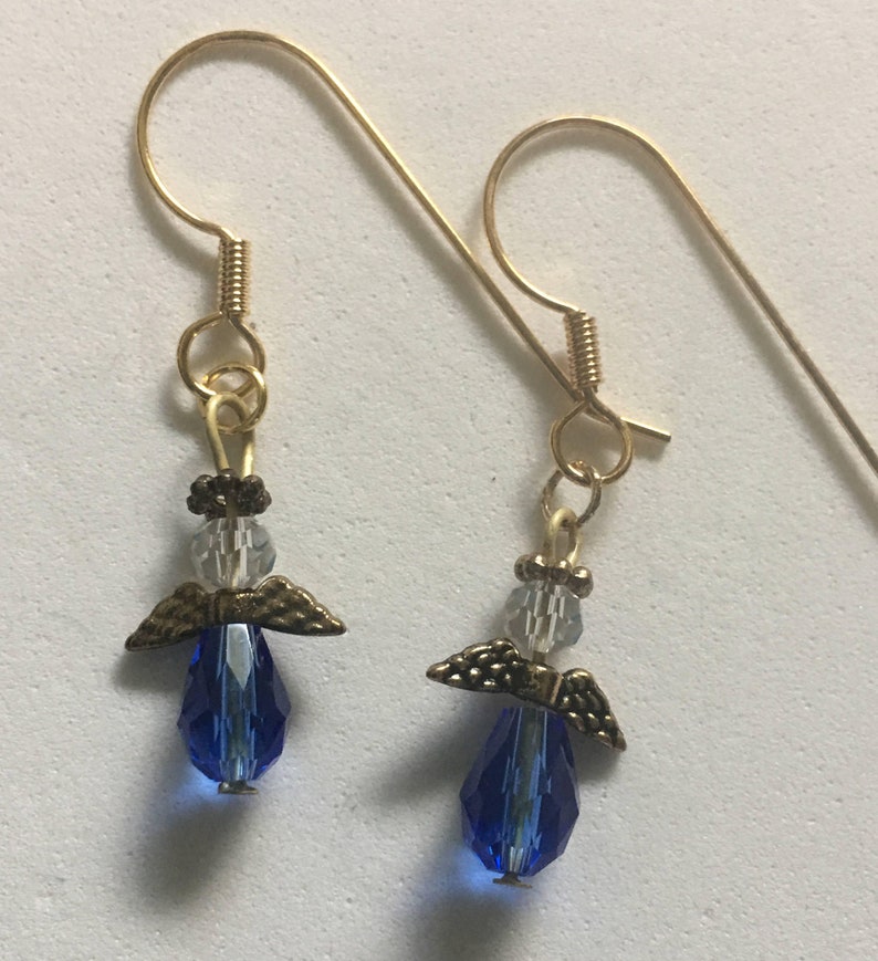 Op de afbeelding: Een paar gouden oorhangers met een blauwe kristallen traan en een klein gouden engelbedeltje met heldere kristallen accenten.