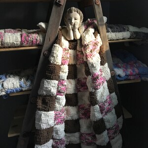 May include: A handmade, checkered blanket in brown, white, and pink hues is draped over a wooden ladder. The blanket's textured design is visible, with other similar blankets on shelves. A small statue of a cherub is placed on the ladder.