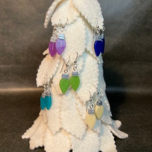 May include: A display of colorful Christmas light bulb earrings. The earrings are in various colors, including purple, blue, green, and yellow. Each earring has a silver-colored cap and hook. The earrings are displayed on a white felt Christmas tree.
