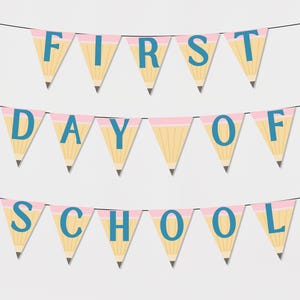 Op de afbeelding: Een slinger met de tekst "FIRST DAY OF SCHOOL" in drie rijen. Elke vlag is ontworpen als een potlood, met een gele behuizing, een roze gum en blauwe letters. De slinger is geregen aan een zwarte draad tegen een witte achtergrond.