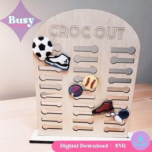 May include: A wooden sign with the text "CROC OUT" and a grid of slots for holding charms. The sign is decorated with various charms, including a soccer ball, a basketball shoe, a baseball, a beach ball, a bowling pin, and a red shoe.