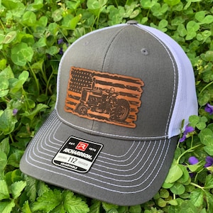 May include: A gray and white trucker hat with a brown leather patch featuring an American flag with a tractor on it. The patch says "Richardson" and "112 Trucker" on it.