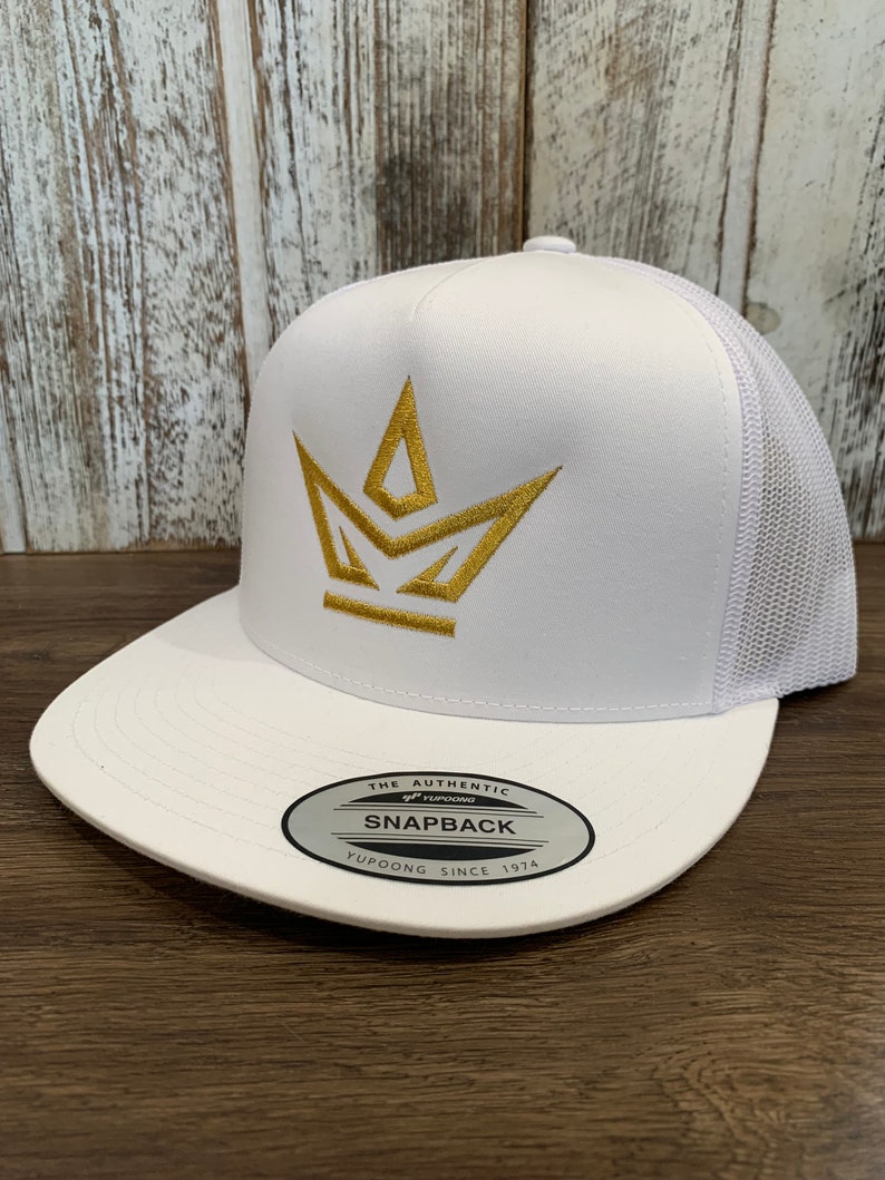 Puede incluir: Gorra de camionero blanca de malla con un logotipo de corona dorada y el texto "THE AUTHENTIC YUPOONG SNAPBACK YUPOONG SINCE 1974" en la etiqueta.