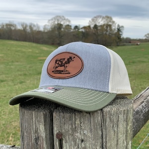 May include: A gray and green trucker hat with a brown leather patch featuring a black and white cow silhouette. The hat has a mesh back.