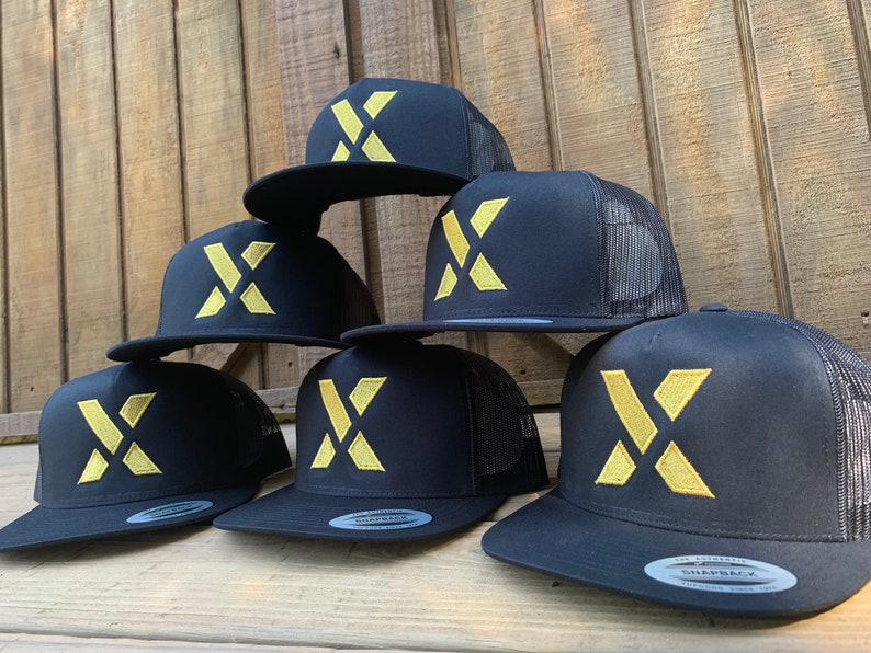 Puede incluir: Seis gorras de camionero negras con un logotipo X bordado en oro. Las gorras est&aacute;n apiladas unas sobre otras.