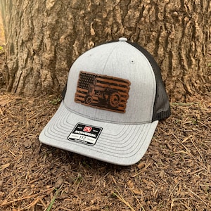 May include: A gray and black trucker hat with a brown leather patch featuring an American flag with a tractor silhouette. The patch has a distressed look. The hat has the text "Richardson 112 Original Trucker Adjustable" on the front.