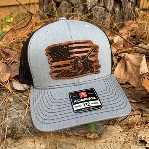 May include: A gray and black trucker hat with a brown leather patch featuring an American flag and a combine harvester. The patch has a distressed look. The hat has the text "Richardson. Original 112 Trucker Adjustable" on the back.