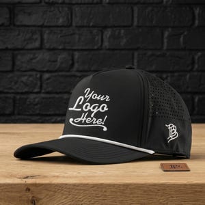 May include: A black baseball cap featuring a white rope detail across the base of the crown and the white embroidered text Your Logo Here on the front. The side panel includes a white monogram logo and perforated ventilation holes for airflow.