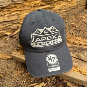 May include: A charcoal gray baseball cap featuring an embroidered white logo of a house roofline with the text APEX ROOFING. A white sticker on the brim displays the number 47, the text 47 CLEAN UP, and the word ADJUSTABLE in black lettering.