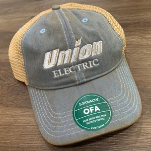 May include: A blue and tan trucker hat with a white embroidered logo that says "Union Electric". The hat has a green circular patch that says "Legacy OFA Visor Board Made From Recycled Content #SPOTTHETAN".