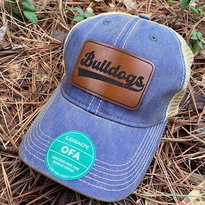 May include: A blue and tan baseball cap with a brown leather patch that says "Bulldogs" in black script. The cap has a small green sticker that says "Legacy OFA Visor Board Made from Recycled Content Let Your Legacy"