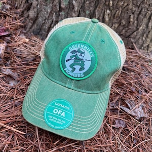 May include: Green and tan baseball cap with the text "GREENHILLS PIONEERS" on a circular patch. The cap has a curved brim and a mesh back. A sticker on the cap reads "OFA" and "VISOR BOARD MADE FROM RECYCLED CONTENT".