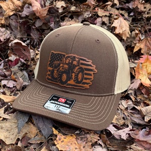 May include: Brown and beige trucker hat with a brown leather patch featuring a tractor silhouette in front of an American flag design. The patch has the text "EST. 1970 RICHARDSON. 112 TRUCKER" on it.