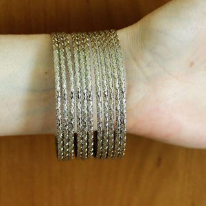 Może przedstawiać: Zestaw srebrnych bransoletek z teksturowanym wzorem, ułożonych na nadgarstku. Bransoletki prezentują skomplikowany design i metaliczny połysk. Każda bransoletka ma szerokość około 0,6 cm, tworząc warstwowy efekt na nadgarstku.