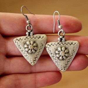 Puede incluir: Un par de pendientes de metal plateado con forma triangular y superficie texturizada. Cada pendiente muestra un motivo floral central en relieve rodeado por un borde en zigzag y está sujeto a un sencillo gancho de metal plateado.
