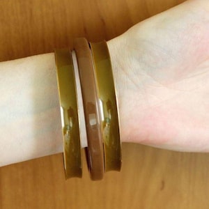 Op de afbeelding: Een set van drie gladde armbanden gedragen om de pols. Twee van de armbanden hebben een glanzende olijfgroene kleur, terwijl de middelste armband een effen mokkabruine kleur heeft. Elke armband heeft een eenvoudig, afgerond ontwerp met een glanzende afwerking.