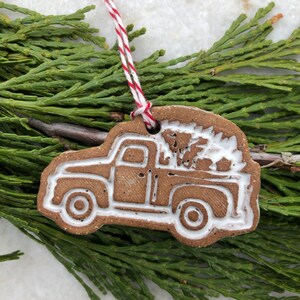 Op de afbeelding: Een bruine keramische kerstversiering in de vorm van een vintage pick-up truck met een witte kerstboom in de laadbak. De versiering heeft een wit touwtje om op te hangen.