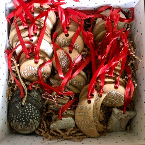 Op de afbeelding: Een doos gevuld met handgemaakte keramische ornamenten. De ornamenten zijn versierd met verschillende ontwerpen, waaronder een kerstboom, een sneeuwpop, een rendier en een kerstman. De ornamenten zijn vastgebonden met rood lint.