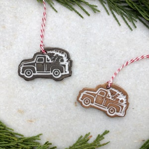 Op de afbeelding: Twee kerstversieringen van keramiek in de vorm van pick-up trucks met kerstboom in de laadbak. De trucks zijn bruin en wit met witte contouren. De ornamenten hangen aan een rood-wit touwtje.
