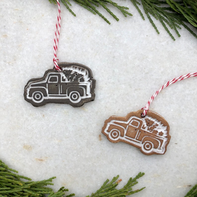 Op de afbeelding: Twee kerstversieringen van keramiek in de vorm van pick-up trucks met kerstboom in de laadbak. De trucks zijn bruin en wit met witte contouren. De ornamenten hangen aan een rood-wit touwtje.