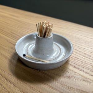 May include: A light gray ceramic match holder with a central cylindrical column for holding matches. The base is a shallow, circular tray with a raised rim. A single wooden match with a dark tip lies horizontally on the tray surface.