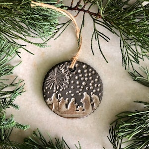 May include: A ceramic Christmas ornament with a brown glaze and a white silhouette of the nativity scene. The ornament features a star and a dotted background.