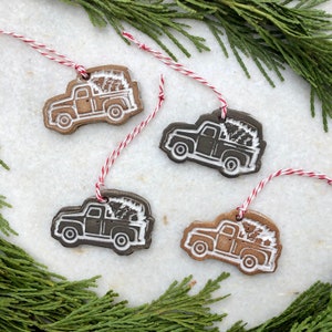 Op de afbeelding: Vier keramische ornamenten in de vorm van pick-up trucks die kerstboom vervoeren. De ornamenten zijn in twee kleurvariaties: bruin met witte details en zwart met witte details. Elk ornament heeft een touwtje om op te hangen.