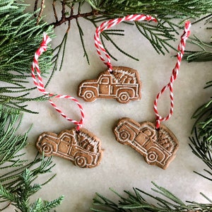 Op de afbeelding: Drie bruine keramische ornamenten in de vorm van pick-up trucks met witte contouren. Elke truck vervoert een kleine kerstboom. De ornamenten hangen aan een rood-wit touwtje.