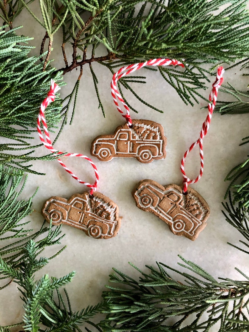 Op de afbeelding: Drie bruine keramische ornamenten in de vorm van pick-up trucks met witte contouren. Elke truck vervoert een kleine kerstboom. De ornamenten hangen aan een rood-wit touwtje.