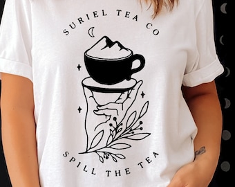 Puede incluir: Camiseta blanca con un gráfico negro de una taza de té con un diseño de montaña, sostenida por una mano con acentos florales. El texto "SURIEL TEA CO" y "SPILL THE TEA" están arriba y abajo del gráfico. La camiseta tiene mangas enrolladas.