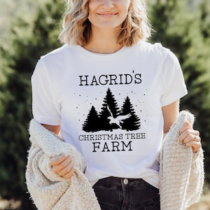 May include: White t-shirt with the text "Hagrid's Christmas Tree Farm" in black. The design includes silhouettes of pine trees and a bird. The shirt is worn by a person with blonde hair, and they are holding a cream-colored sweater.