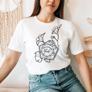 May include: White t-shirt with a black and white illustration of a chocolate frog card from the Harry Potter series. The card features a frog sitting on a leaf with the words "Chocolate Frog" written on it.