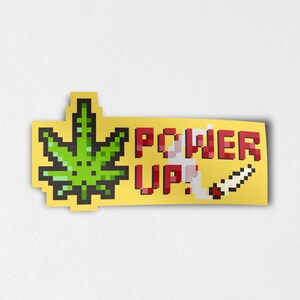 以下が含まれることがあります： ピクセル化された大麻の葉と「POWER UP！」という赤い文字と白い文字が描かれた黄色のステッカー。 燃えているジョイントのピクセル化された画像がステッカーの右側にあります。