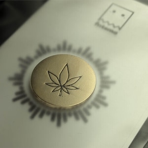 May include: A circular gold-toned metal pin featuring an engraved seven-pointed leaf design. The pin is attached to a white rectangular card displaying a black graphic of a stylised ghost-like figure and a radiating sunburst pattern behind the pin.